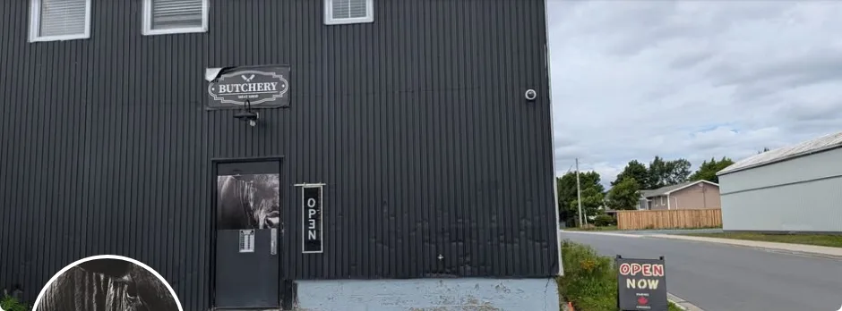 Black corrugated storefront of The Butchery with its Butchery sign over the door and an open sign beside it, Bay Roberts